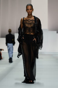 Model walks the runway in a sheer black ensemble: lace crop top, flowing sheer pants, and a matching robe with white piping.