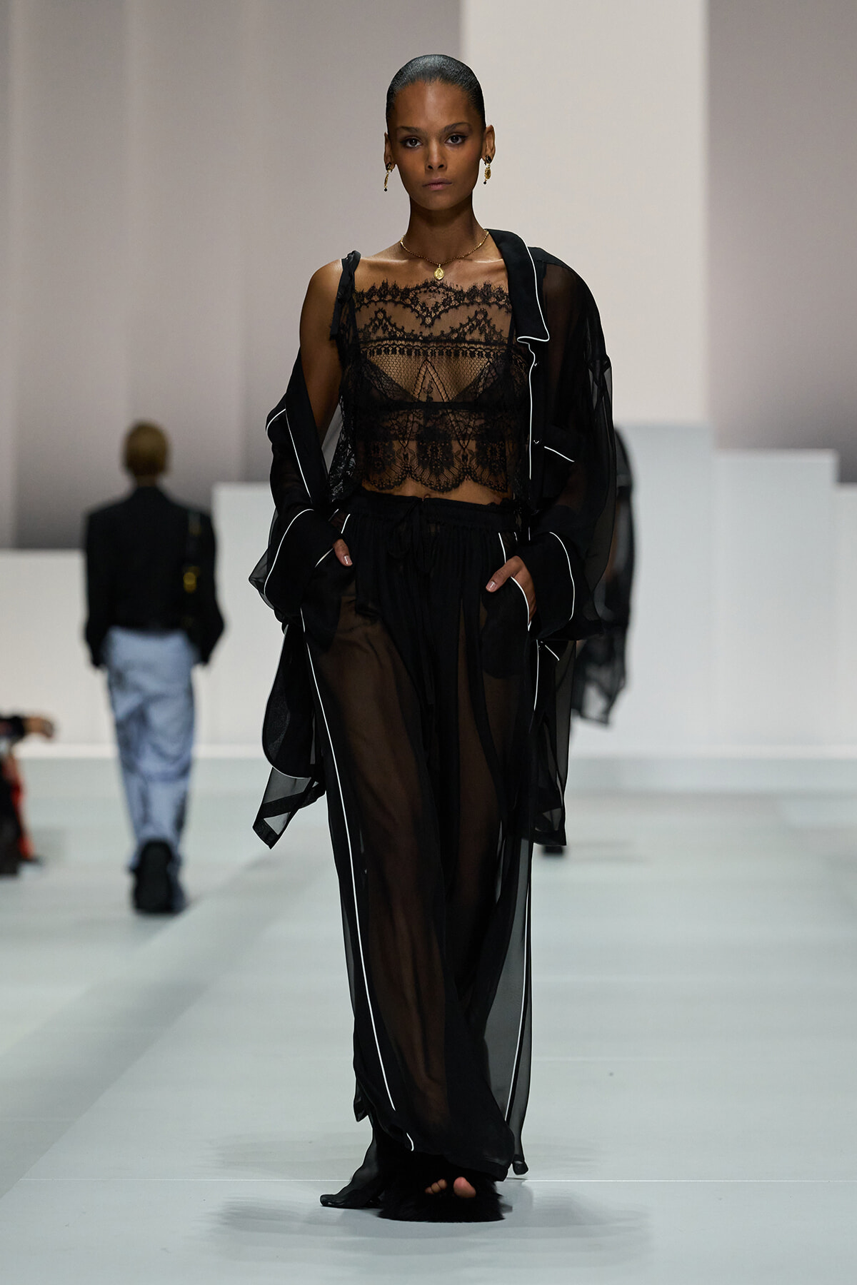 Model walks the runway in a sheer black ensemble: lace crop top, flowing sheer pants, and a matching robe with white piping.