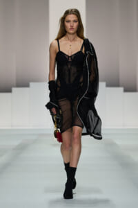 Runway model walks in a sheer black dress with a structured bodice and black boots, holding a clutch with a red tassel
