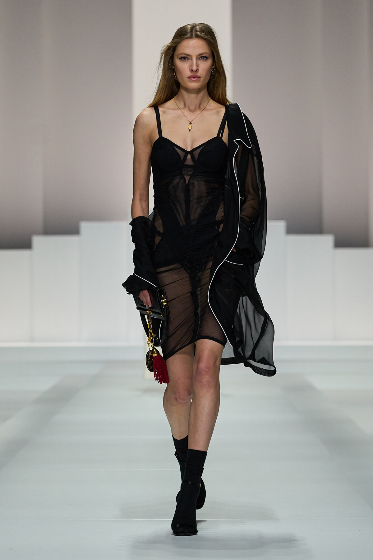 Runway model walks in a sheer black dress with a structured bodice and black boots, holding a clutch with a red tassel