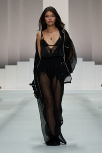 Model walks the runway in a black sheer robe over a lace-trimmed black lingerie top and sheer pants, accented with a pendant necklace on a minimalist set.