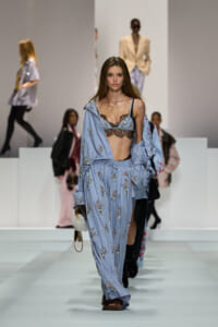 Model walks the runway in a blue striped, embroidered pajama-style outfit with a lace bralette and coordinating shirt.