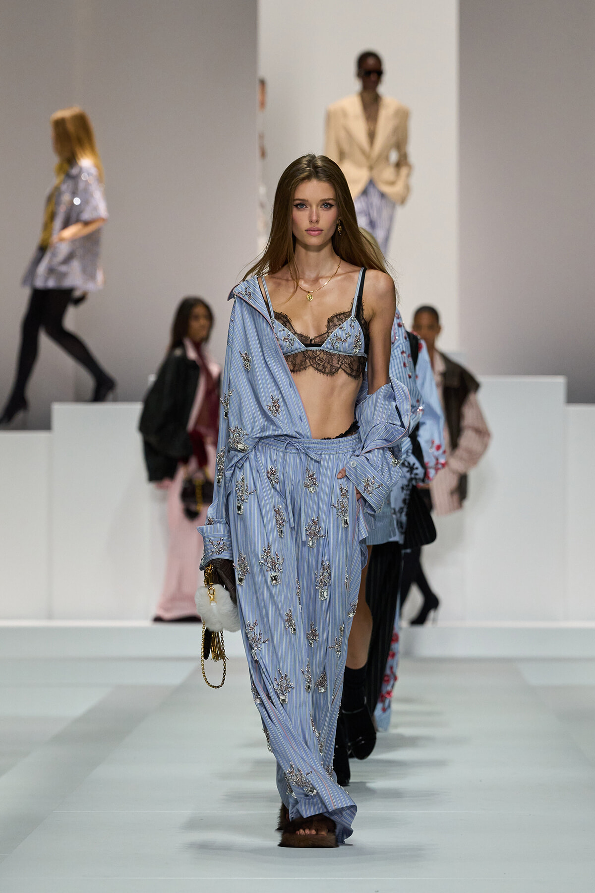 Model walks the runway in a blue striped, embroidered pajama-style outfit with a lace bralette and coordinating shirt.
