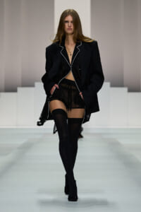 Model on a runway wearing an oversized black blazer with white piping, sheer black lingerie, thigh-high stockings, and black ankle boots, with a cross necklace.