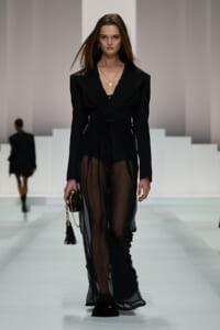 Model walking a runway in a black tailored blazer with a sheer, wide-leg overlay and belt; gold pendant necklace and small black clutch with a tassel.