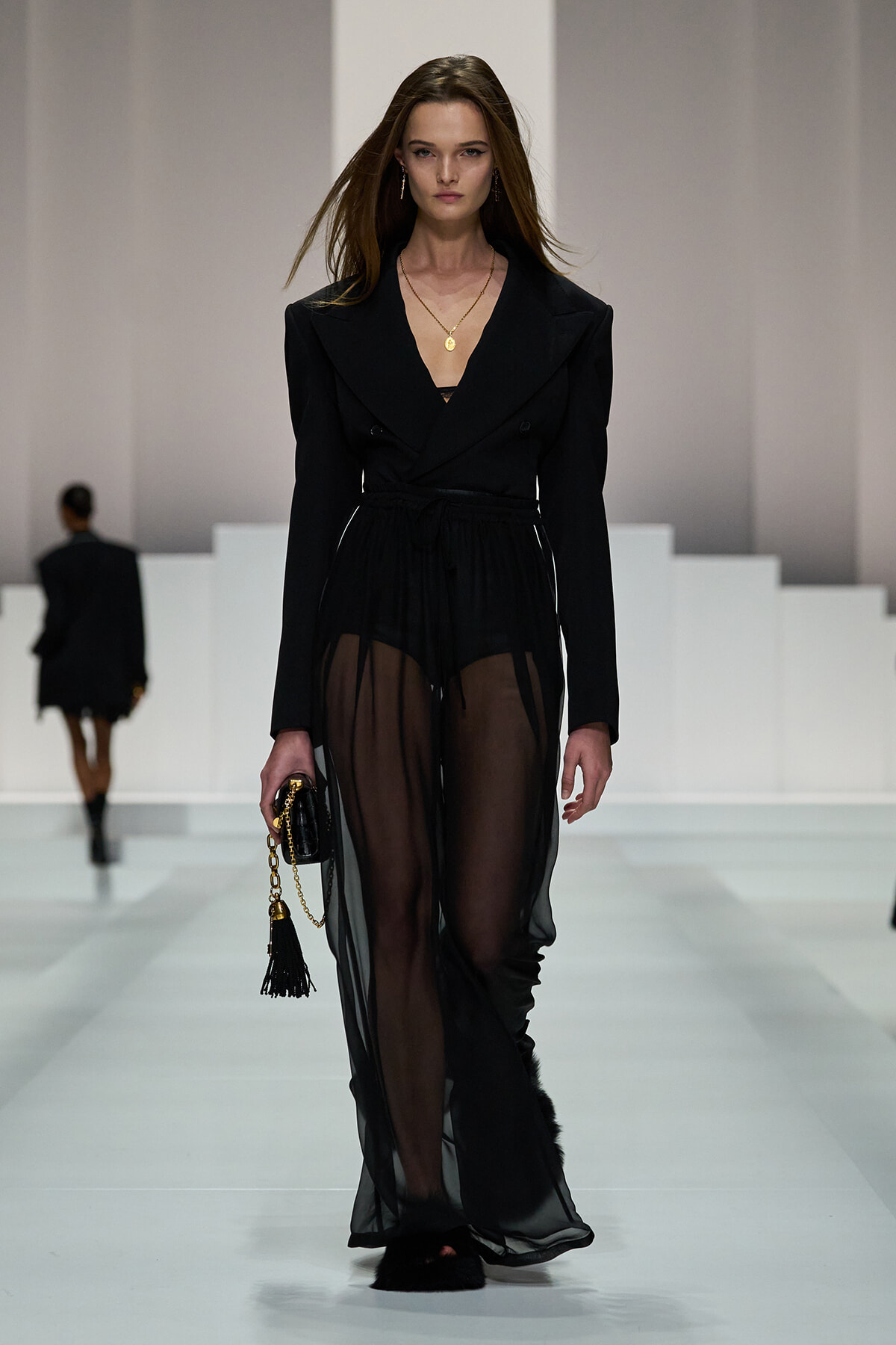 Model walking a runway in a black tailored blazer with a sheer, wide-leg overlay and belt; gold pendant necklace and small black clutch with a tassel.