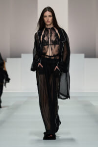 Model walking a runway in a black sheer lace top with matching sheer robe and wide-leg pants, hands in pockets.