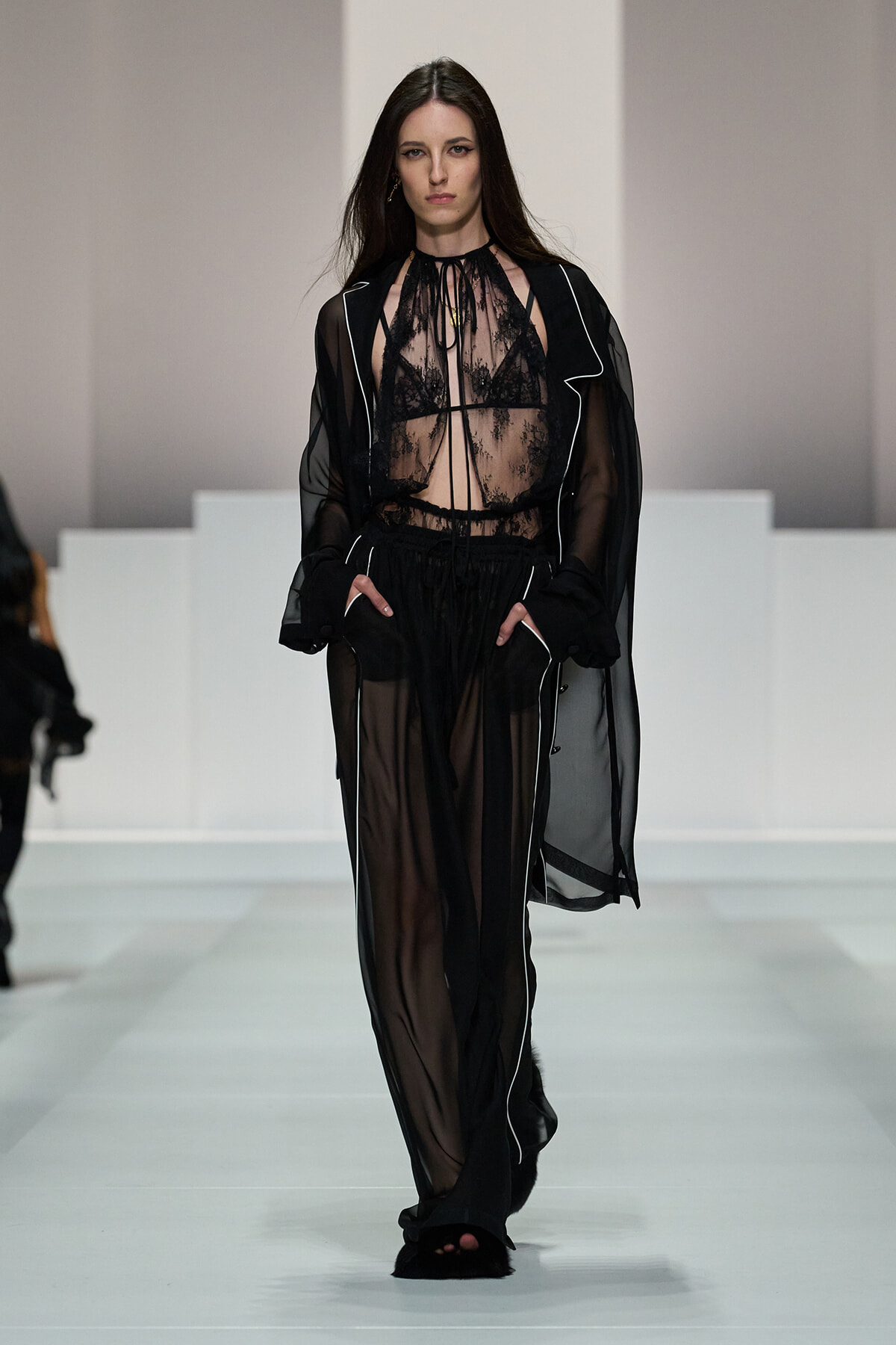Model walking a runway in a black sheer lace top with matching sheer robe and wide-leg pants, hands in pockets.