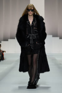 Model walking a runway in a long black fur coat over a sheer black outfit with white piping and a waist tie, wearing dark sunglasses.