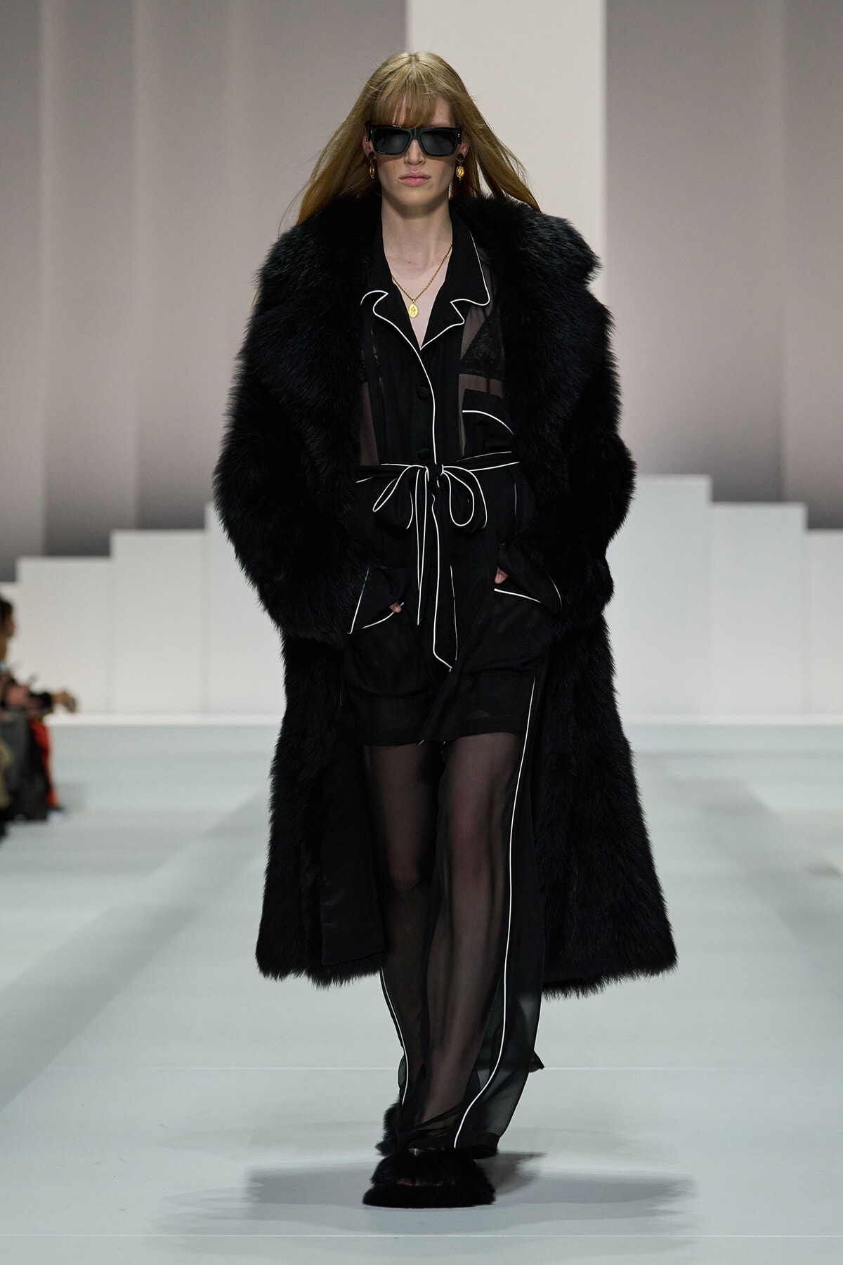 Model walking a runway in a long black fur coat over a sheer black outfit with white piping and a waist tie, wearing dark sunglasses.