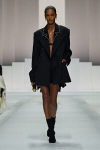 Runway model wearing an oversized black blazer with white piping, open at the front, paired with black ankle boots.