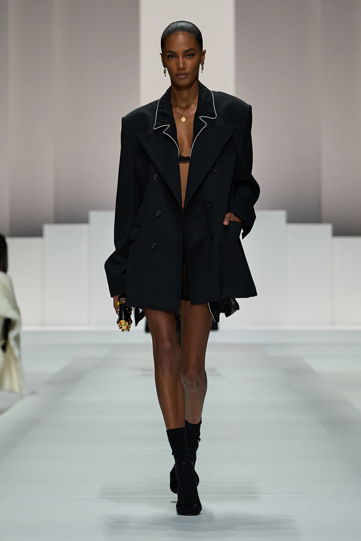 Runway model wearing an oversized black blazer with white piping, open at the front, paired with black ankle boots.