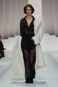 Model walks a fashion runway in a sheer black jumpsuit with white piping, paired with a white fur coat draped over one shoulder.