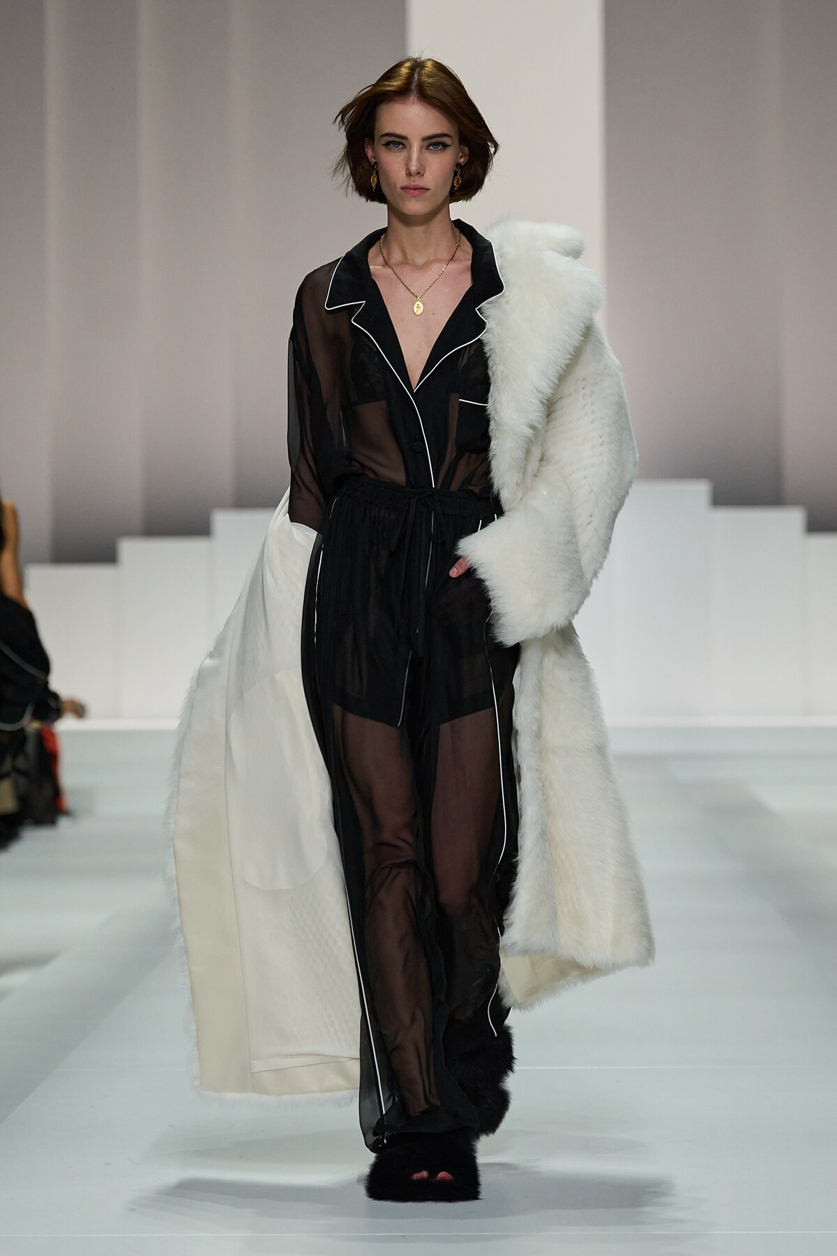 Model walks a fashion runway in a sheer black jumpsuit with white piping, paired with a white fur coat draped over one shoulder.