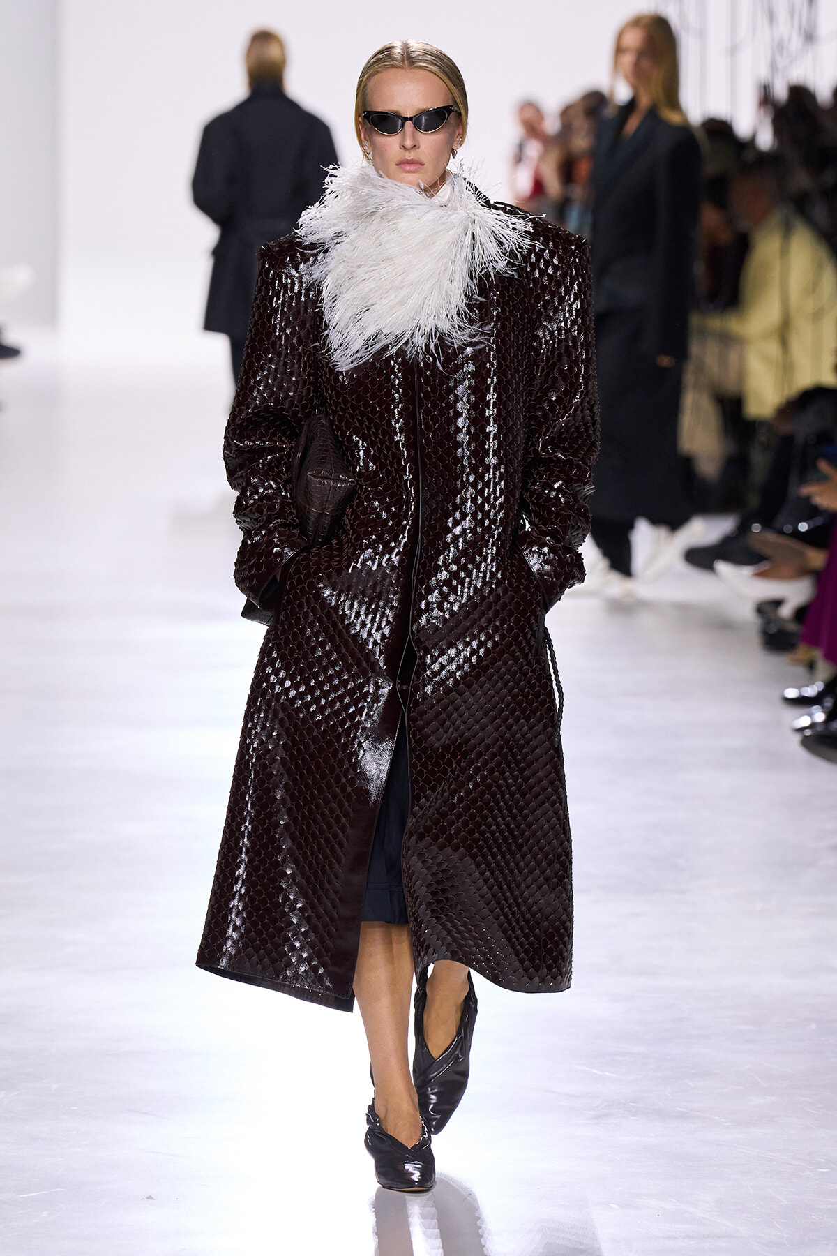 Model on a runway wearing a long, glossy, snakeskin-textured coat with a white feather scarf and dark sunglasses.
