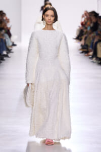 Runway model in a full-length white, textured, fluffy-looking dress with long sleeves and pink slide sandals, carrying a white fluffy clutch.