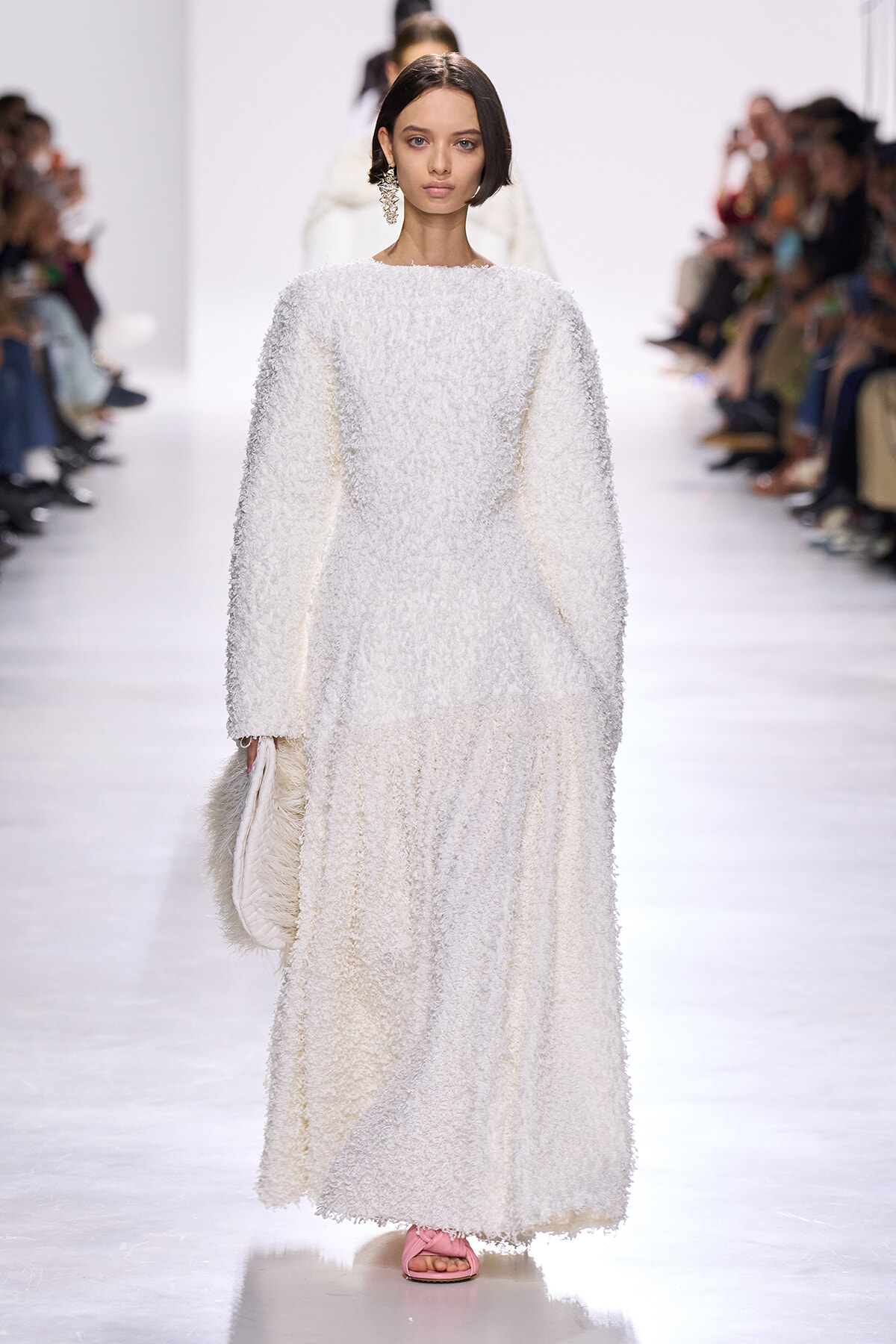 Runway model in a full-length white, textured, fluffy-looking dress with long sleeves and pink slide sandals, carrying a white fluffy clutch.