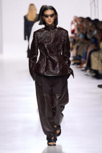 Female model walking a runway in a glossy dark brown textured top and oversized brown pants with sunglasses.