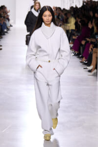 Model walks the runway in a white quilted oversized coat and matching pants with a white belt, hands in pockets, yellow shoes visible.
