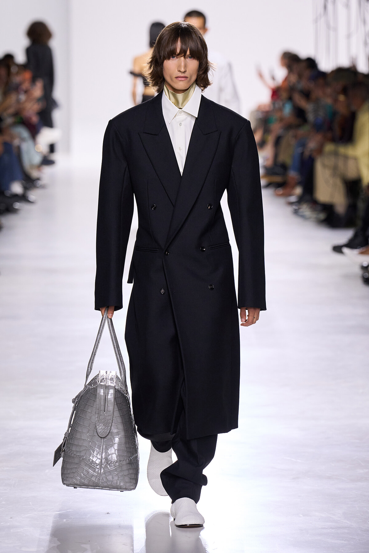 Model on a fashion runway wearing a long black double-breasted coat with a white shirt, carrying a large silver crocodile-embossed bag.