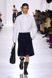 Model walking a runway in a crisp white button-down shirt tucked into a navy textured midi skirt, black boots, and carrying a black tote bag.