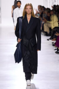 Fashion model walking a runway in a black coat with fringe details, carrying a textured blue handbag.