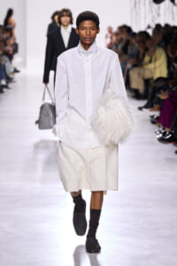Male model on a runway wearing a white button-down shirt and knee-length cream shorts, holding a large white fluffy bag.