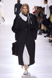 Model on a runway wearing a black oversized trench with a braided belt, white quilted scarf, and white inner layers while carrying a dark croc-embossed bag.