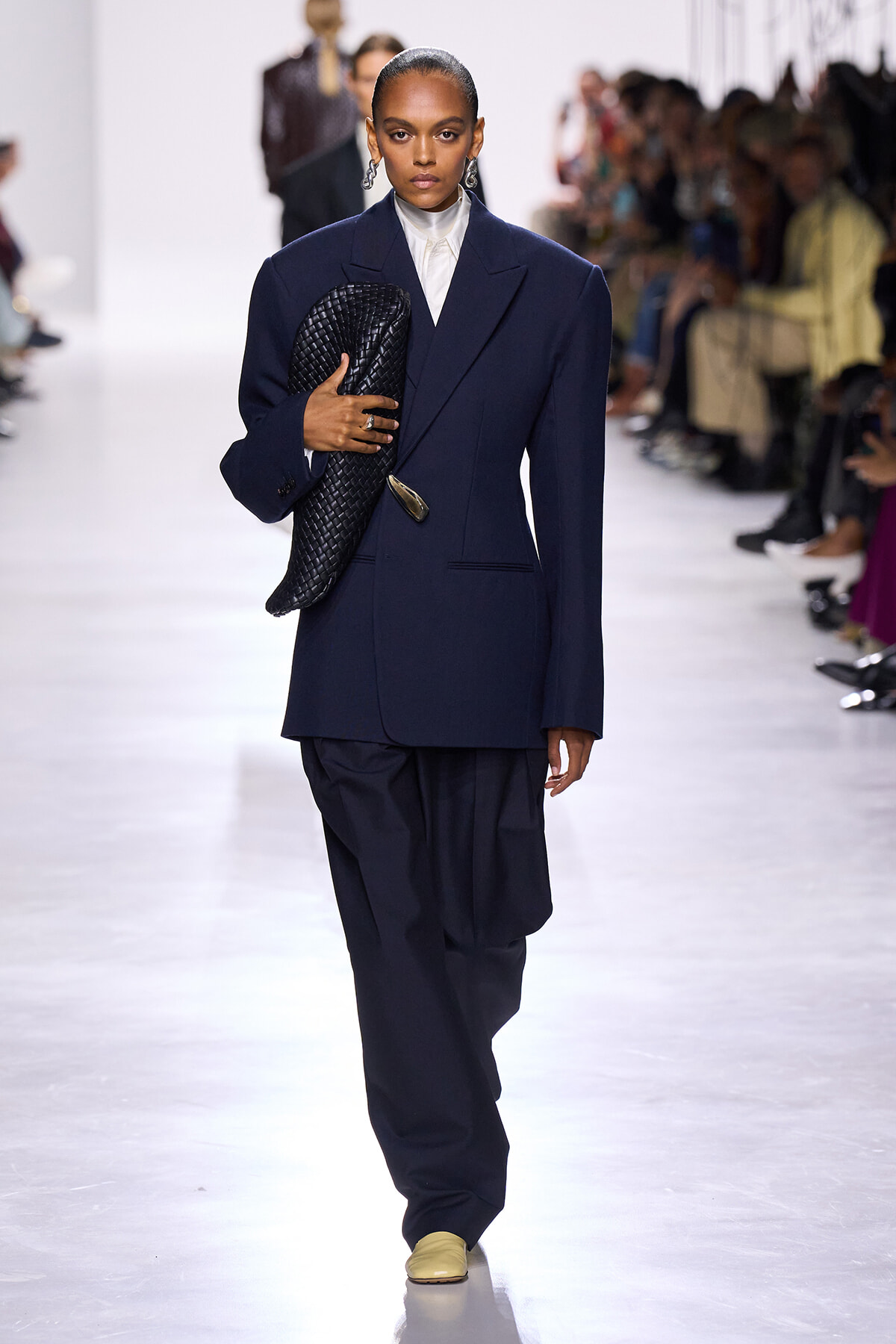 Model walks the runway in a navy oversized suit with a white shirt, holding a black woven clutch under her arm.