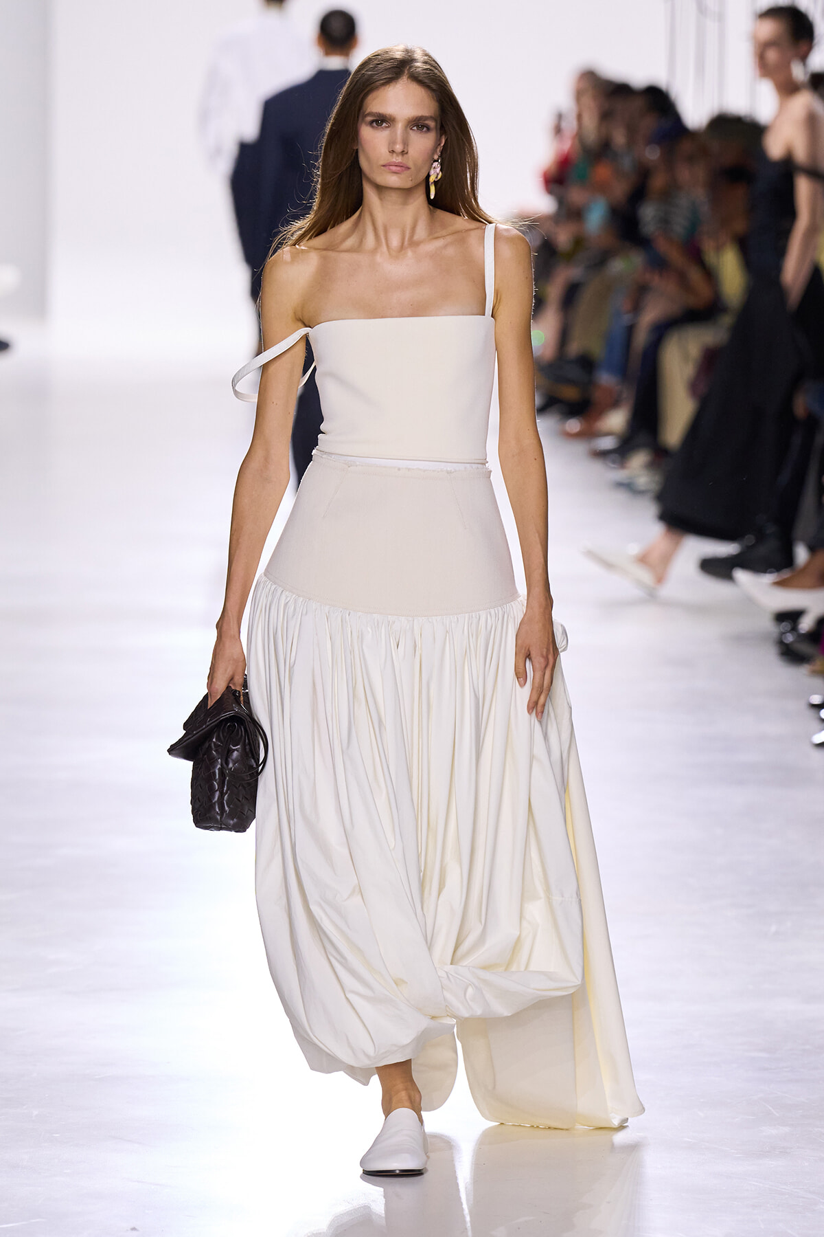 Fashion model walking a white, one-shoulder top and full, flowing skirt on a runway, carrying a black quilted handbag.