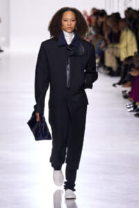 Model walking a fashion runway in a long black coat with a navy collar and front toggle, paired with dark pants, a white turtleneck, and white sneakers; carrying a dark blue quilted clutch as spectators watch.