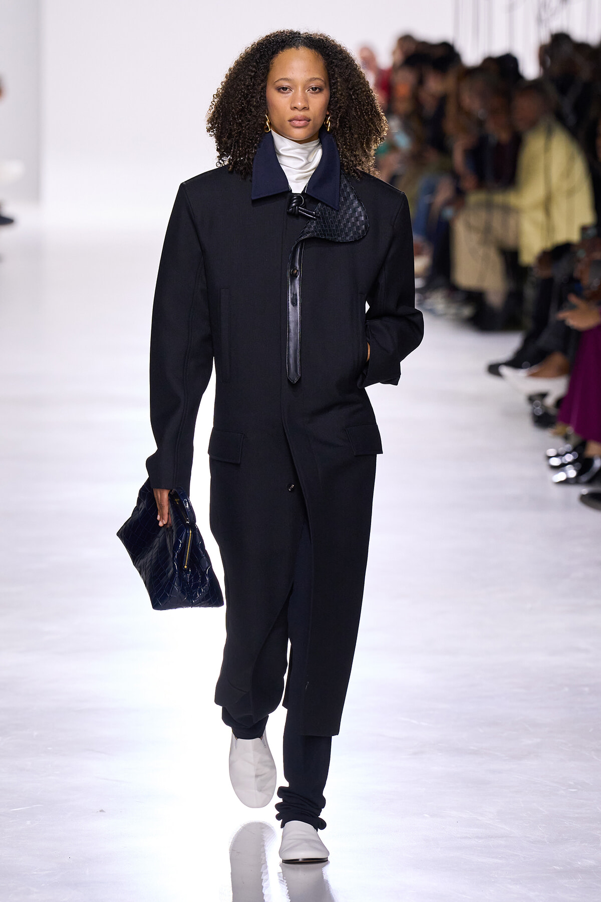 Model walking a fashion runway in a long black coat with a navy collar and front toggle, paired with dark pants, a white turtleneck, and white sneakers; carrying a dark blue quilted clutch as spectators watch.