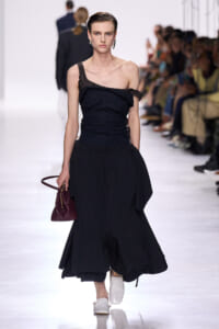 Model walks the runway in a black asymmetric top with a ruched bodice and a voluminous black skirt; she holds a small burgundy handbag and wears white shoes.