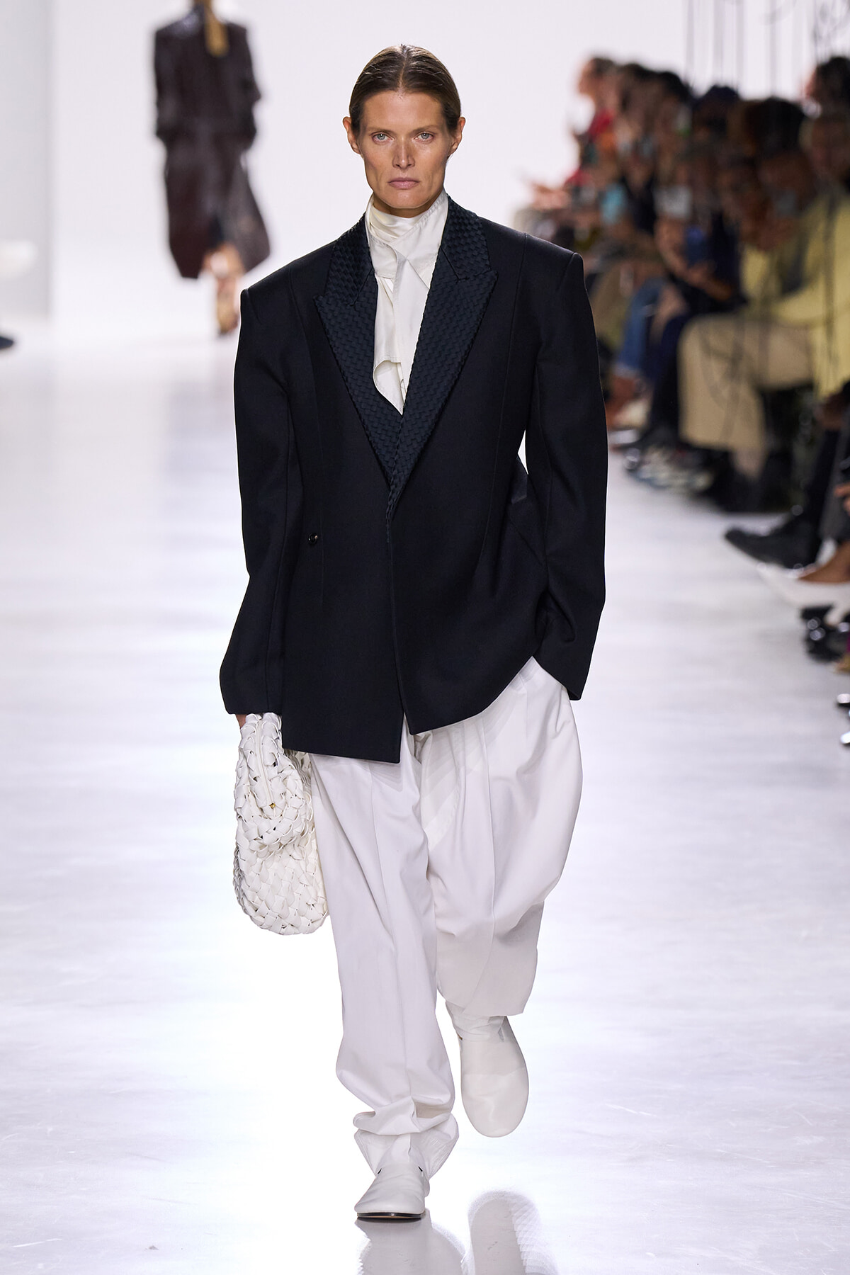 Model walking runway in oversized black blazer with white ruffled blouse and white wide trousers, carrying a white woven bag.