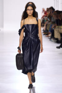 Model walking a dark blue leather dress on a runway, carrying a quilted black bag and wearing black shoes and glasses.