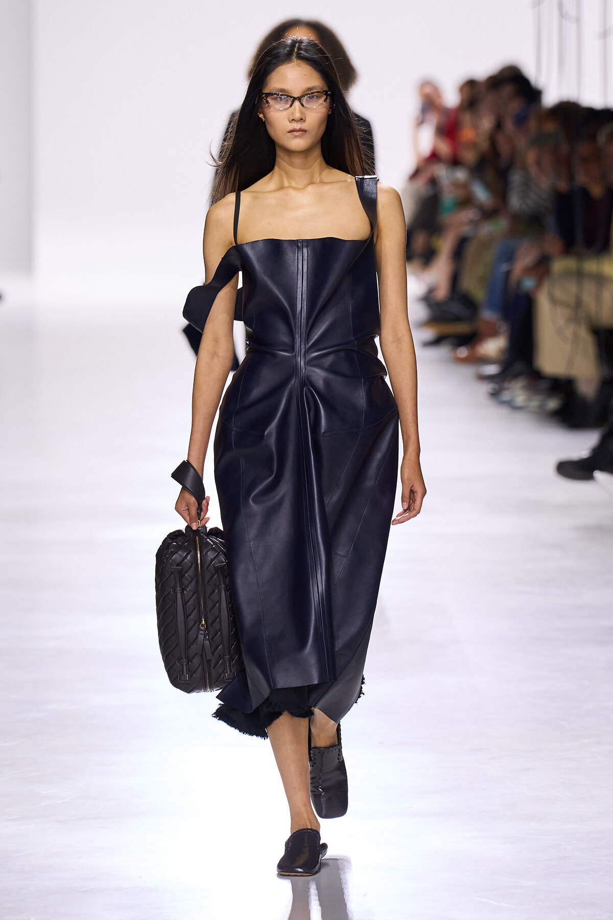 Model walking a dark blue leather dress on a runway, carrying a quilted black bag and wearing black shoes and glasses.