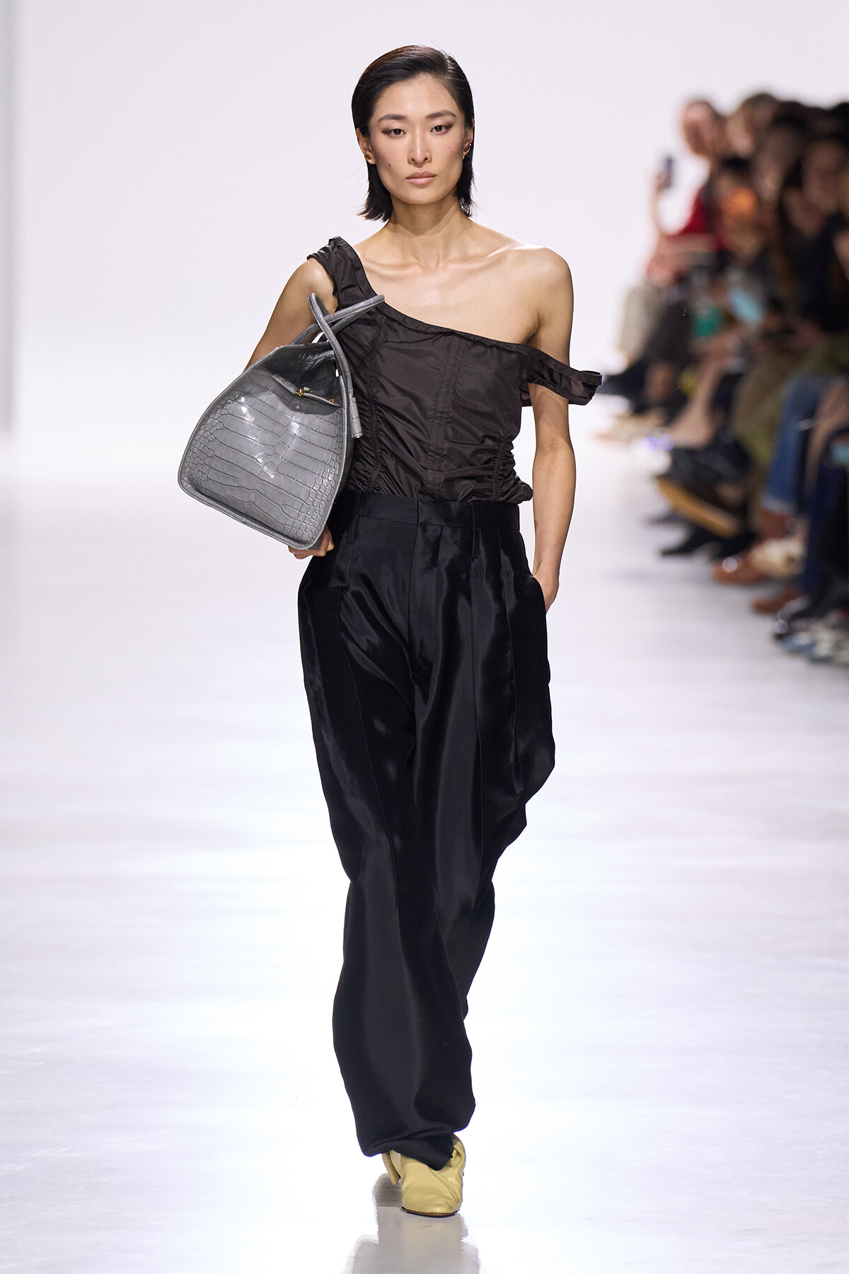 Model walks a fashion runway in an off-shoulder black top and black satin wide-leg pants, carrying a gray croc-embossed handbag on her left.