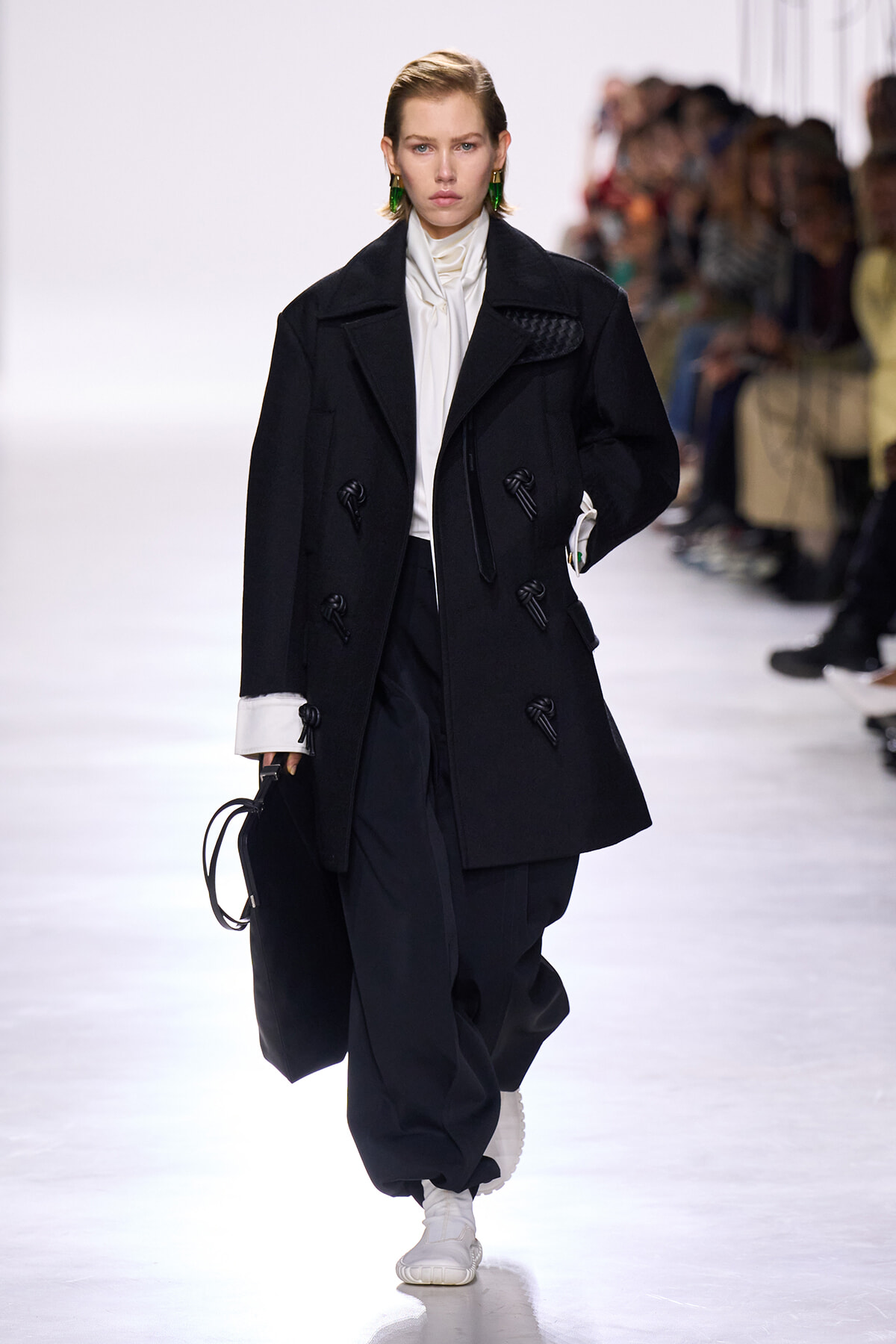 Model on a runway wearing a long black coat over a white blouse, black trousers, and green statement earrings, carrying a black bag.