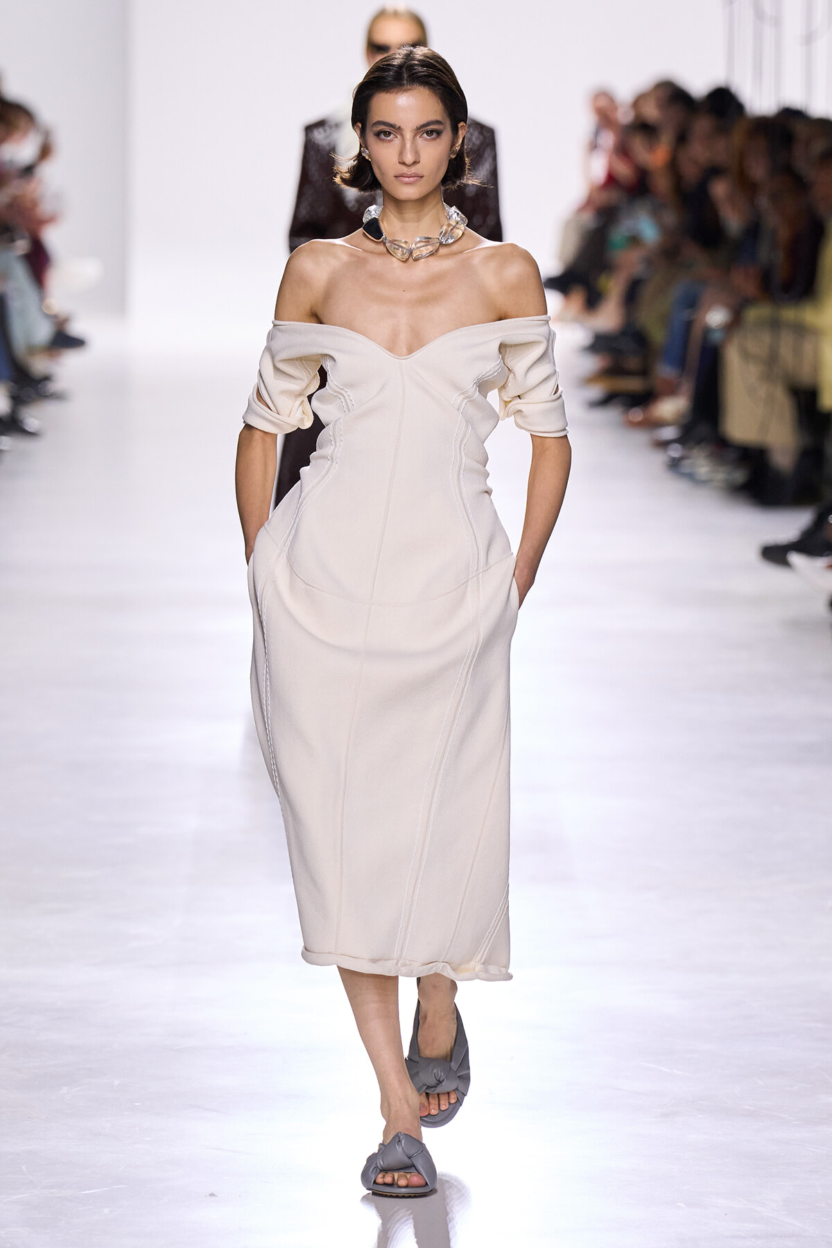 Runway model in a cream off-shoulder midi dress with sculpted seams, hands in pockets, and a chunky necklace.