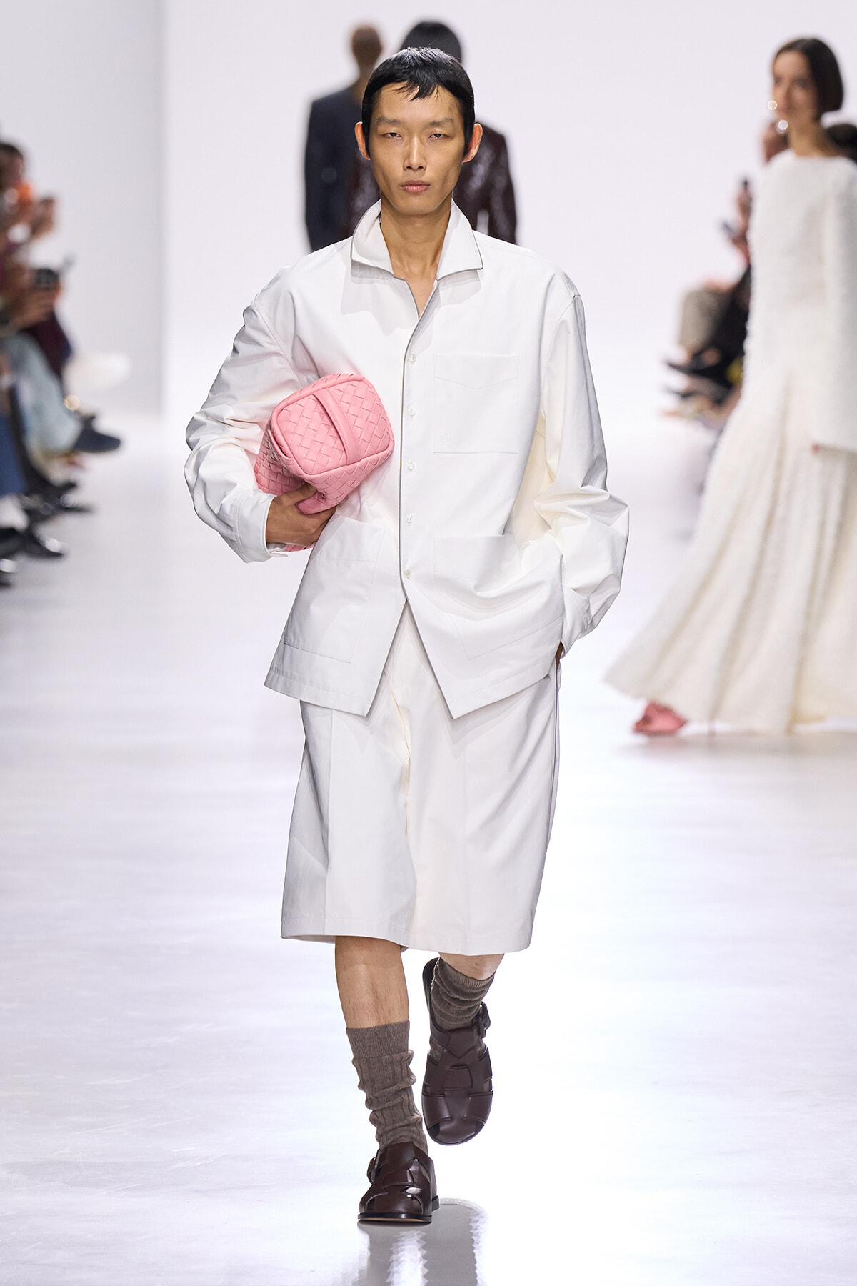 Male model in an all-white oversized shirt and knee-length shorts walks a runway, holding a pink quilted clutch under his arm.