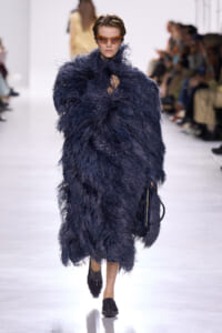 Model walking a runway in a voluminous navy feathered coat with orange-tinted sunglasses and black shoes.