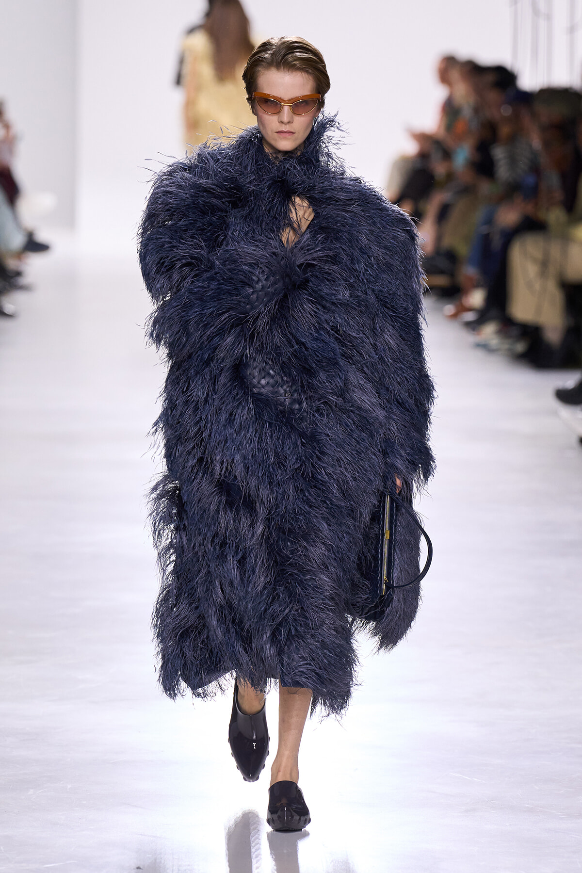 Model walking a runway in a voluminous navy feathered coat with orange-tinted sunglasses and black shoes.