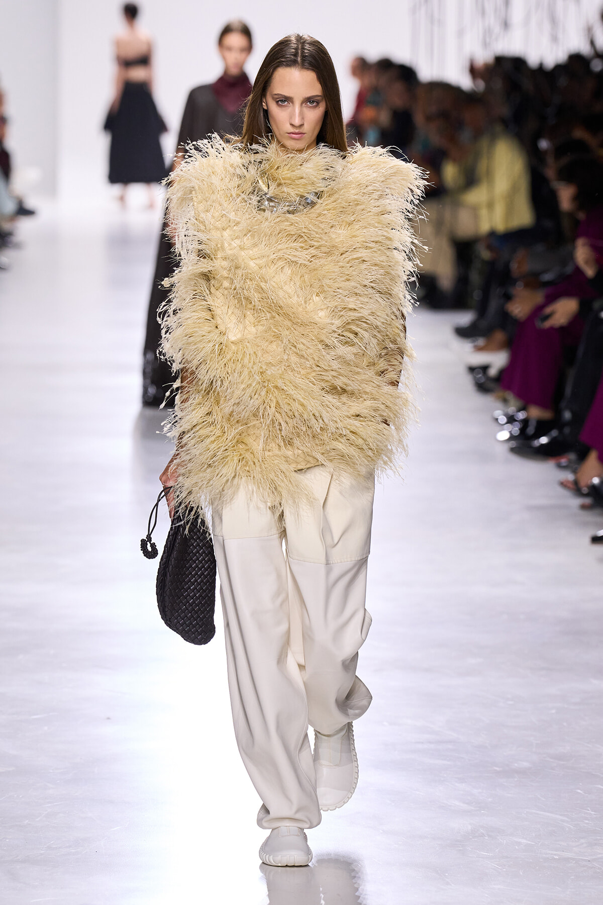 Model walks a runway in a beige feathered top with wide sleeves, paired with cream cargo-style pants and white sneakers, carrying a black quilted handbag.