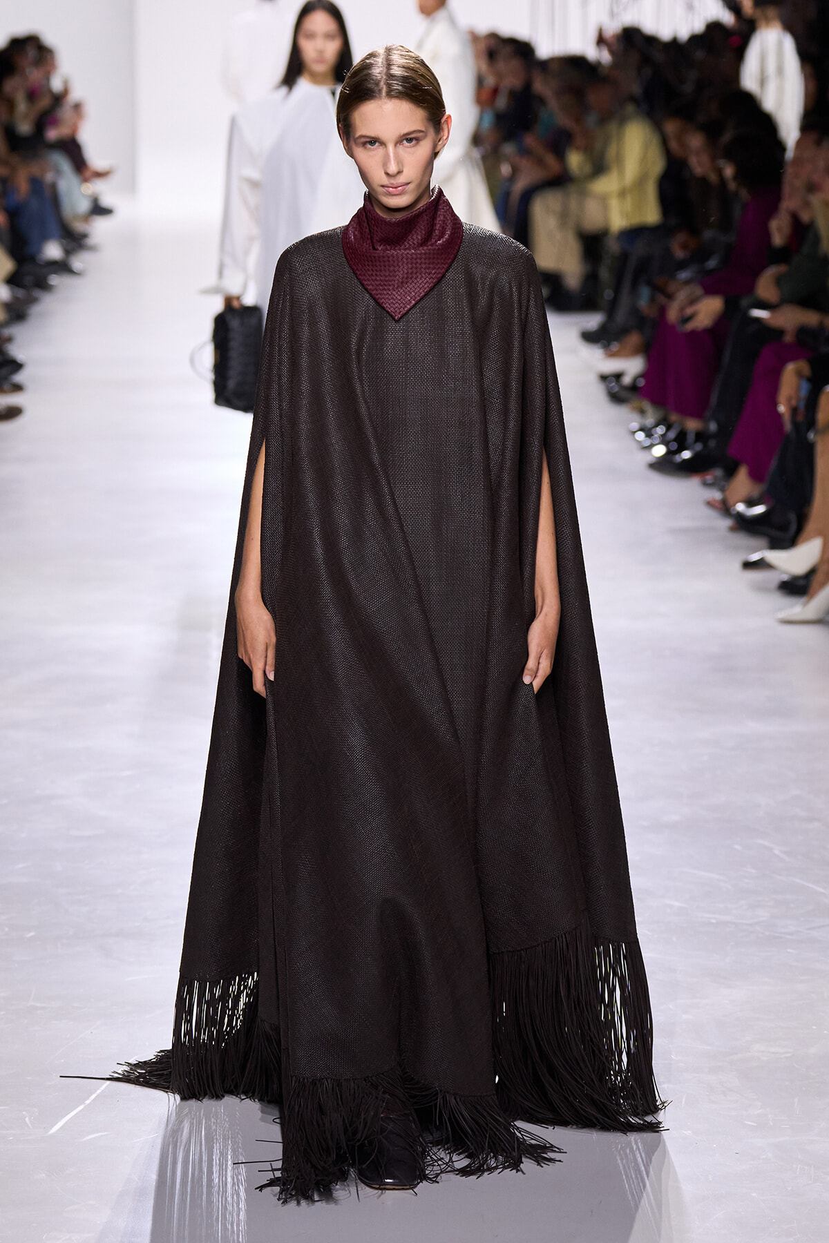 Model walks the runway in a long dark cape with fringe hem, paired with a burgundy turtleneck, during a fashion show.