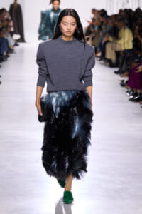 Model walking a fashion show in a gray knit sweater and a voluminous blue‑black fur-like skirt, with bright green shoes.
