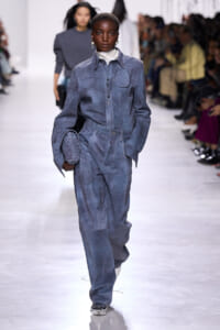 Model walks a fashion runway in a blue oversized shirt-jacket and wide-leg pants set, white turtleneck, and a quilted blue clutch; audience blurred in background.