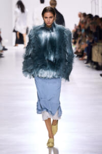 Model walks the runway in a teal feathered top and light blue skirt at a fashion show.