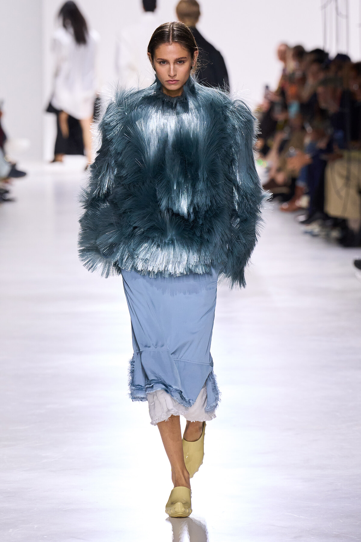 Model walks the runway in a teal feathered top and light blue skirt at a fashion show.