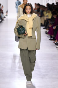 Model wearing a loose beige suit with matching scarf, holding a dark green quilted handbag on a runway