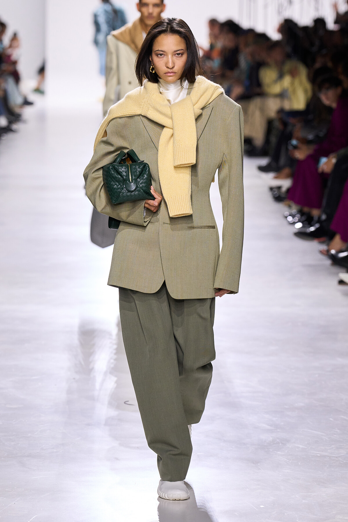 Model wearing a loose beige suit with matching scarf, holding a dark green quilted handbag on a runway